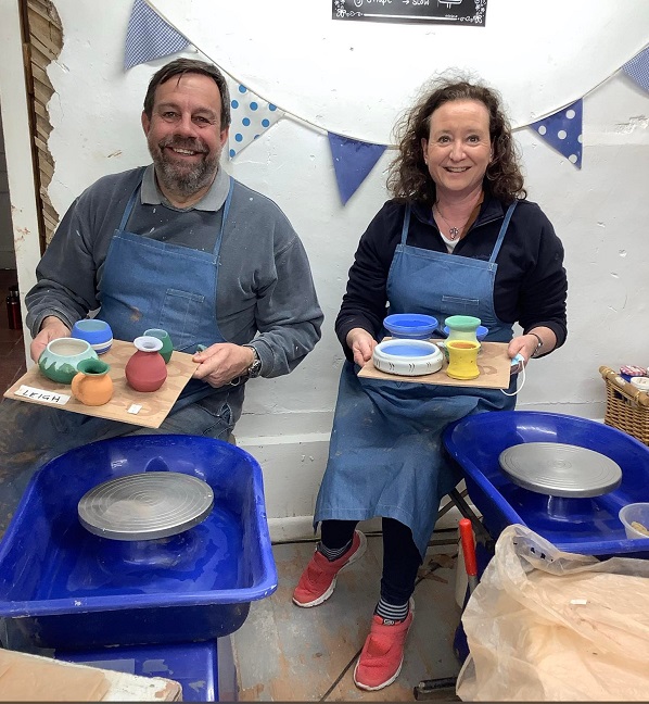 husband and wife smiling sat at potters wheel at eastnor pottery herefordshire holding a board each of painted pots