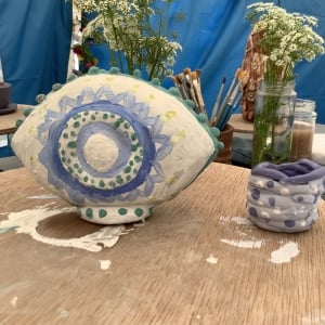 clay modeled eye sculpture painted white and purple with a small coil pot painted purple aside