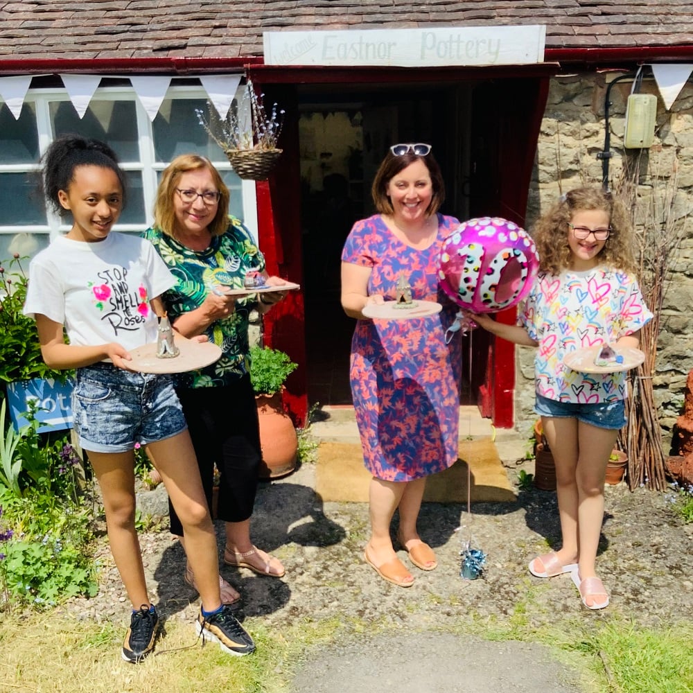 2 adults and 2 children stood outside pottery holiding 10th birthday baloon and trays with their clay creations made at eastnor pottery