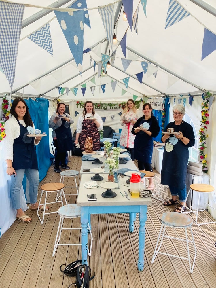 6 women standing round a table inside a marquee wearing aprons and holding wooden boards with hand modeled clay