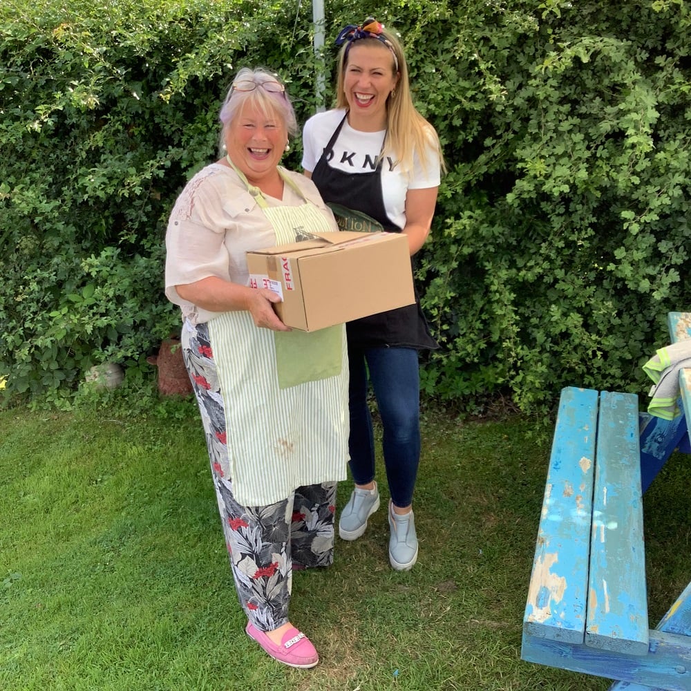 mother and daughter holding cardboard box smiling, in front of hedgerow