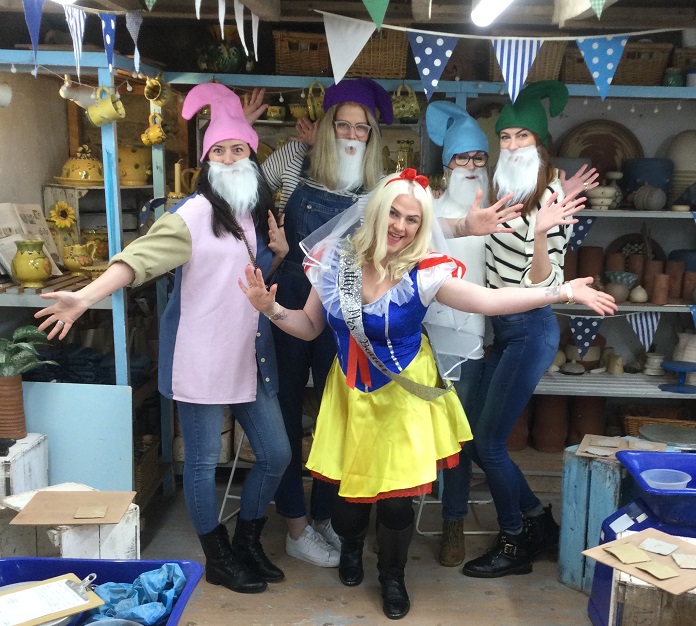 Five ladies stood in the studio at Eastnor Pottery, four are wearing elf hats and beards, one lady in the middle is dressed as Snow White. They have their arms out wide and are smiling and having fun!