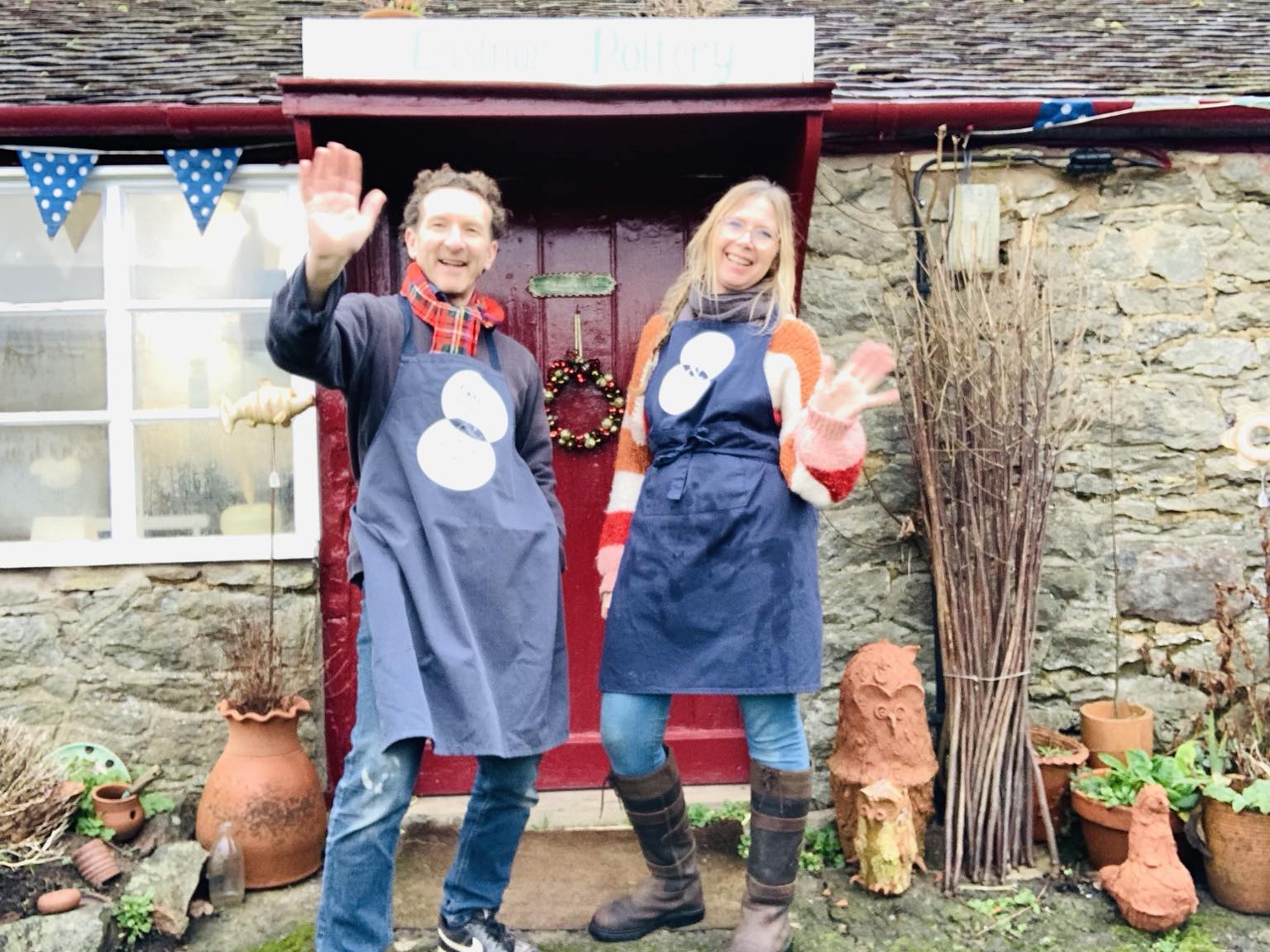 eastnor pottery owners jon williams and sarah monk stood outside eastnor pottery in blue aprons waving at the camera 20th december 2024