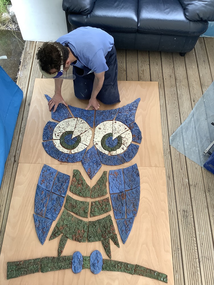 potter jon williams leaning over putting blue white and green tiles hand modeled with clay which together make up an owl 