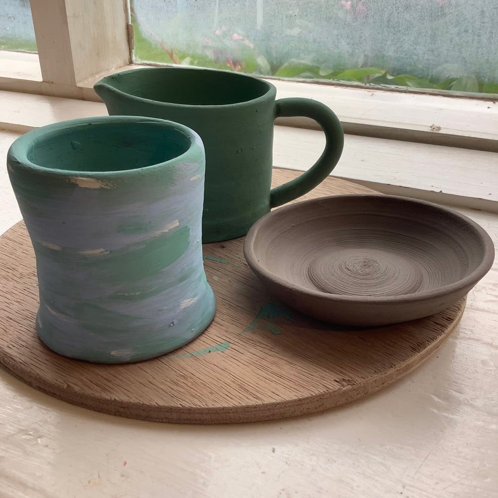 a green jug, green and purple pot and an unpainted ceramic dish on a wooden board