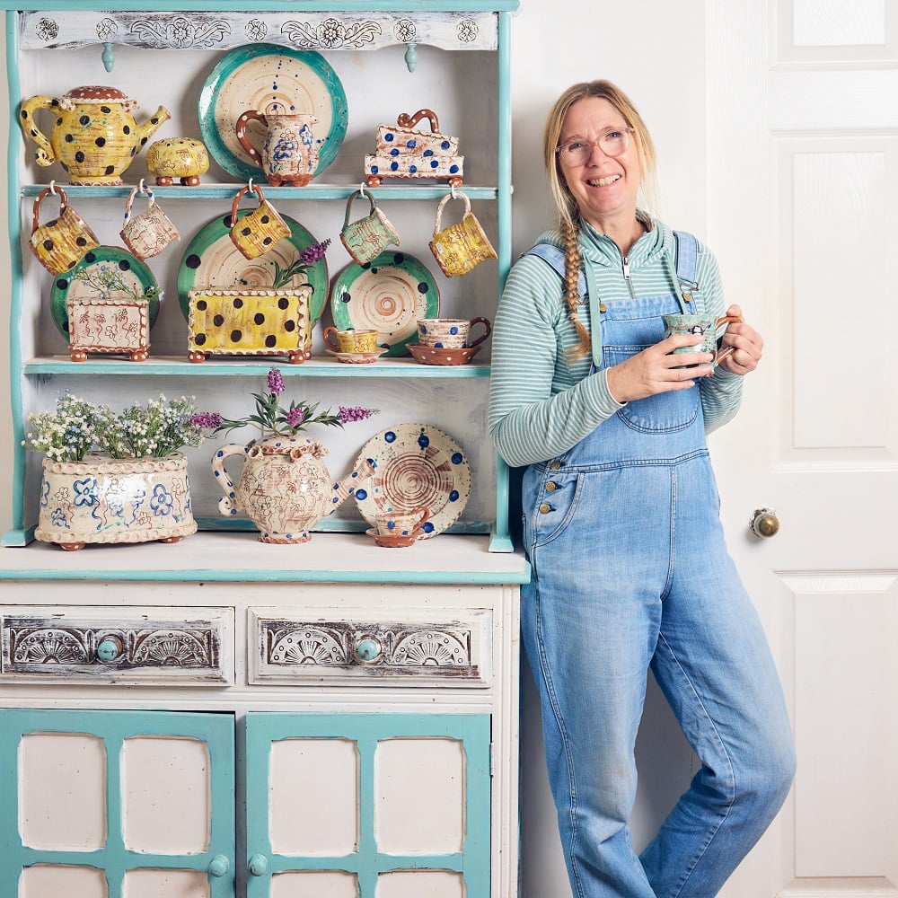 Sarah Monk potter, stood in here studio at Eastnor Pottery. She is leaning against a dresser full of her colourful slipware pottery. She is holding one of her hand thrown mugs and is smiling to the camera.