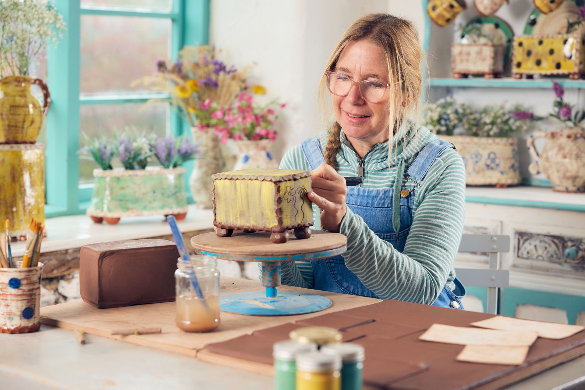 Slipware potter, Sarah Monk sat at he workshop table making a clay flower brick. She is sat by the window, surrounded by her own unique pottery designs.