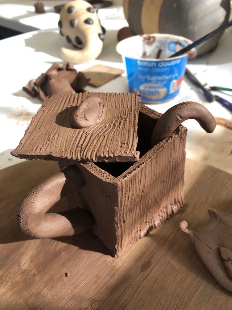 a cubed clay box made from soft clay. it has a lid resting on the top set a jaunty angle to show the box interior. the box has a handle made from clay and a protruding clay spoon handle from inside the box