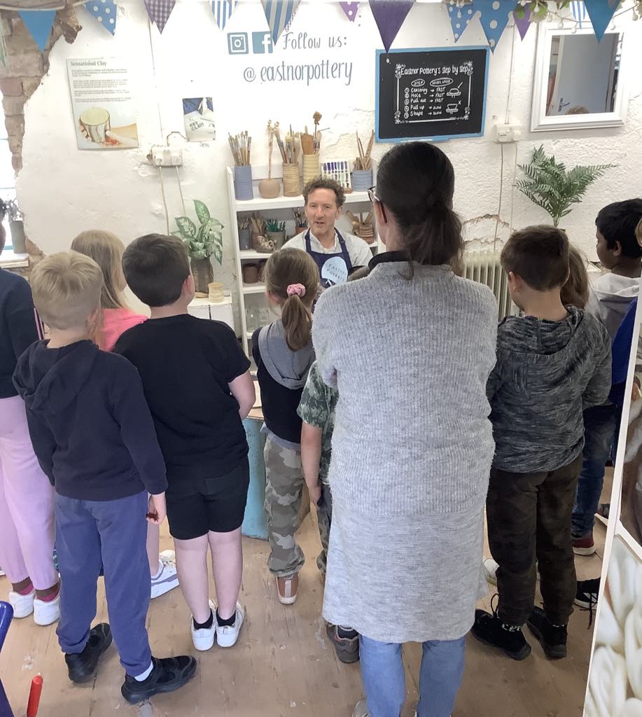 crowd of 19 children and staff from brockhampton primary school watching jon williams from eastnor pottery throw a pot on the potter's wheel. Jon is facing the camera, the children and adults have their backs to the camera