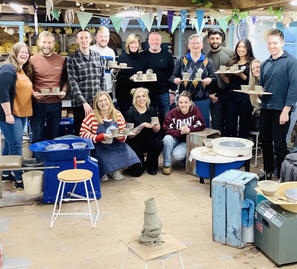 14 very happy class participants holding their pots on boards and looking forward to adding to the re-cycling tower of pots in the center of the pottery studio.