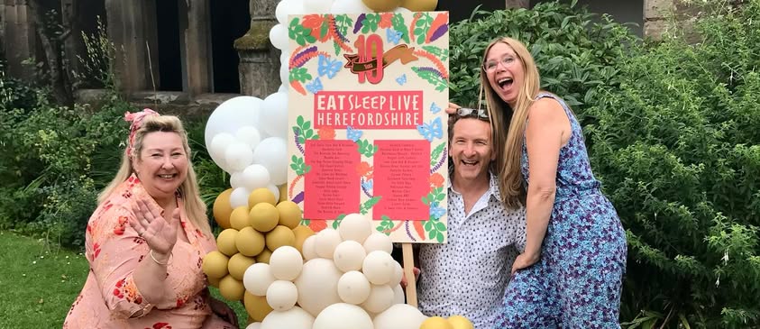 Three smiling people in the garden of Hereford Cathedral. Gathering around a colourful display of balloons and a display board showing a list of gold award winners.
