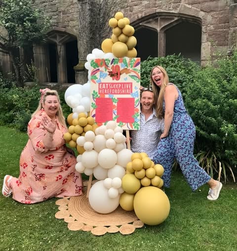Eat Sleep Live Herefordshire balloon display around an easel containing a list of gold award winners. Three happy people are gathered around kneeling on the grass and laughing.