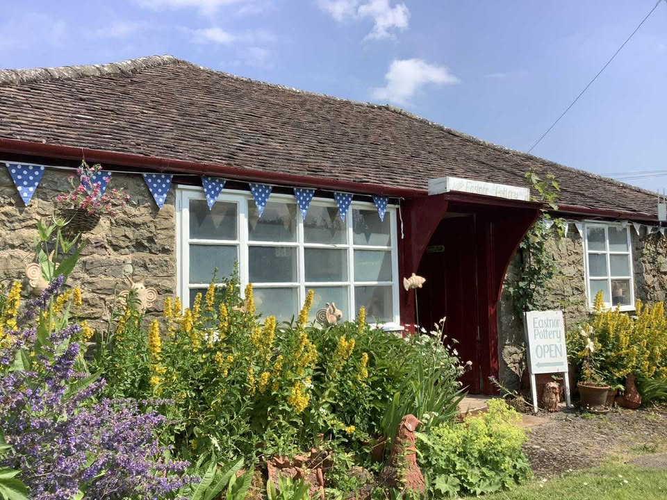 Eastnor Pottery entrance door, on a sunny spring day. Each side of the door there's a cottage garden with flower borders full of yellow and purple flowers. The door is open ready to welcome visitors. The doorway is painted red with an over hanging wooden porch roofing. There is a notice board outside which read 'Eastnor Pottery Open'. Blue and white polka dot bunting hangs each side of the doorway
