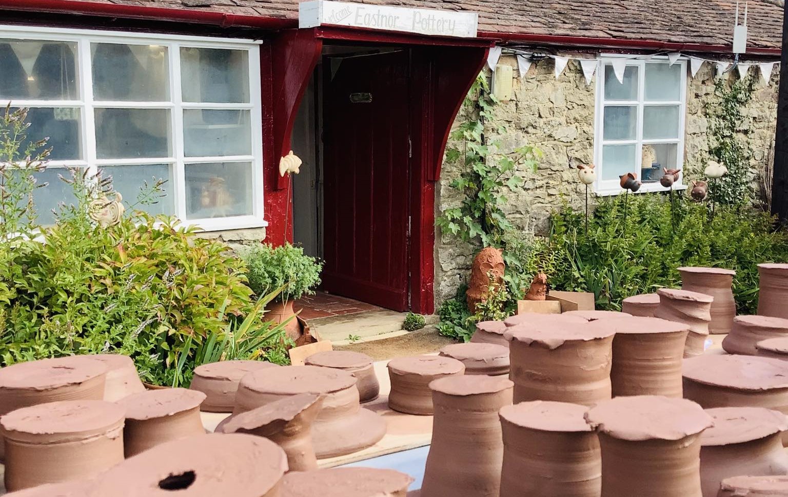 Outside shot of Eastnor Pottery frontage, red front door with white windows frames, garden borders and customer pots turned upside down and drying on tables.