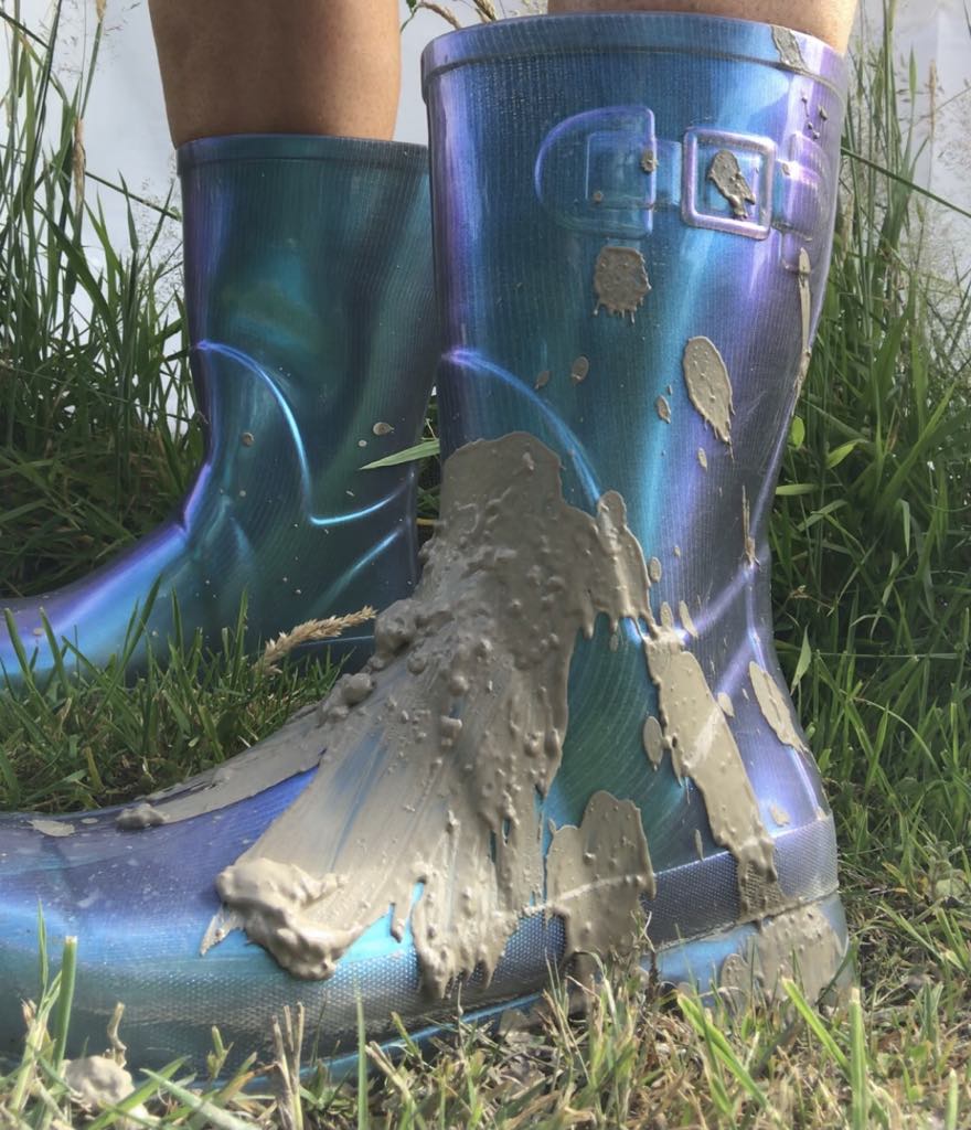A close up of someone wearing some bright blue festival wellies with a splatter of grey mud across them. They are stood on grass.