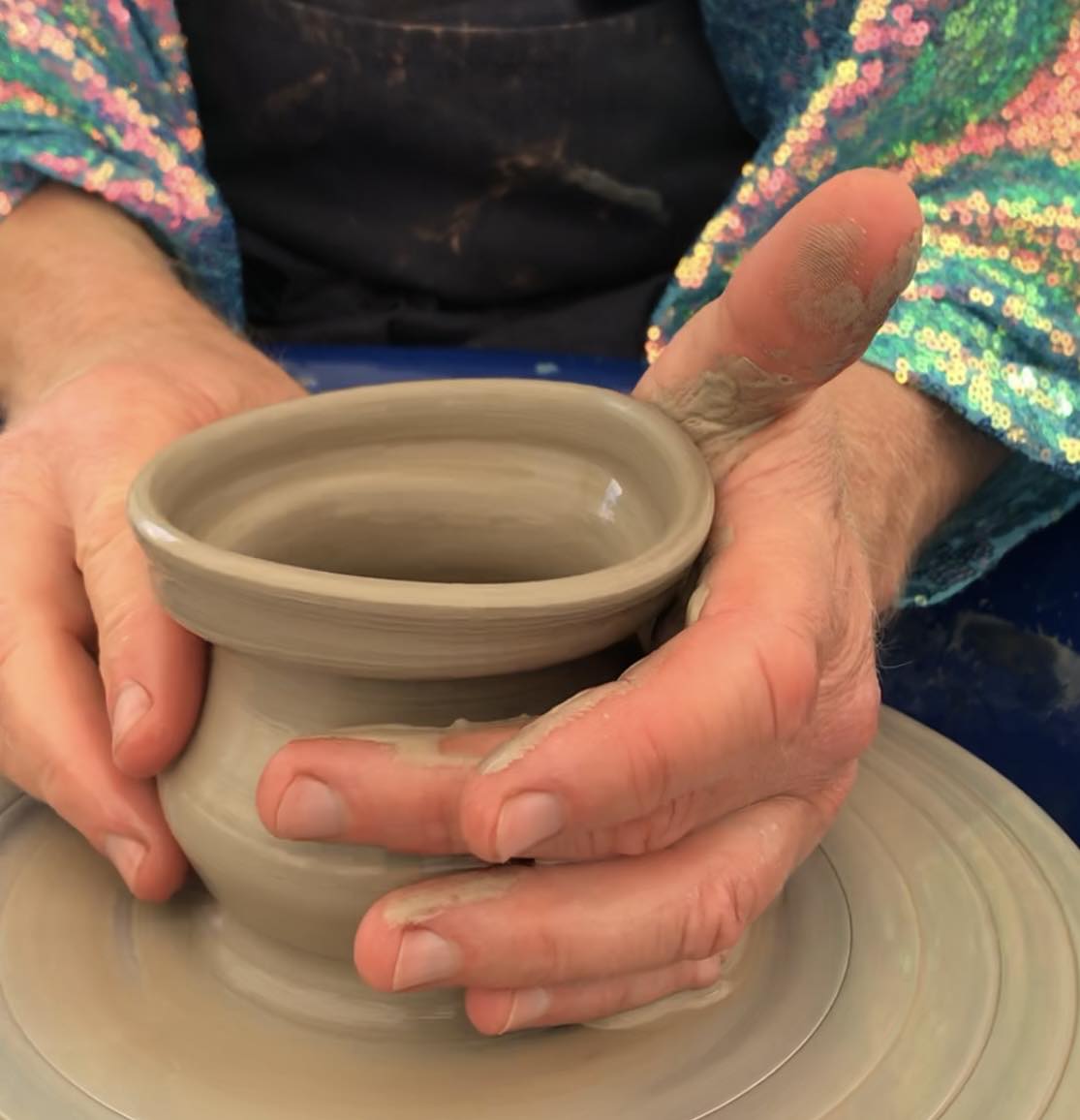 Hands cupped around a wet, freshly made pot on a potter's wheel. The person is wearing sequin sleeves of pink, gold and light blue.