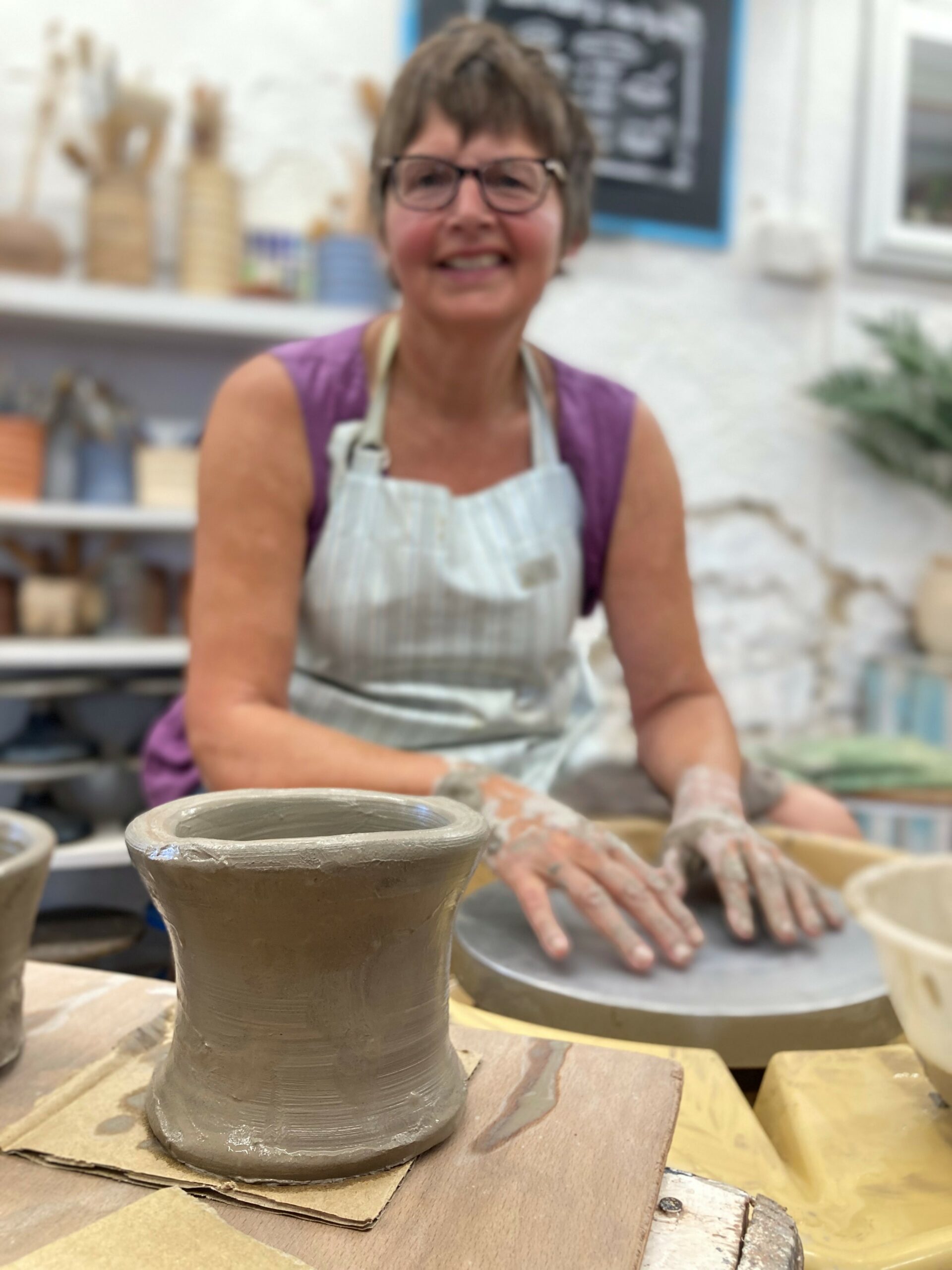 A portrait shot of class participant Gilly. She is sat at a potter's wheel in the studio at Eastnor Pottery. She is wearing a stripey apron and is smiling contentedly. Gilly's hands are resting on the wheel head and she is looking into the camera. Her hands are covered in grey clay. To the for front of the photo is Gilly's freshly thrown pot. A tall cylinder that flutes out  at the top slightly.