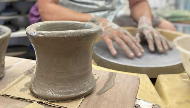 Wet freshly made clay pot on a board in front of a potter's wheel with Gilly's hands resting on the wheel head. Her hands are covered in grey clay, the pot is grey too.