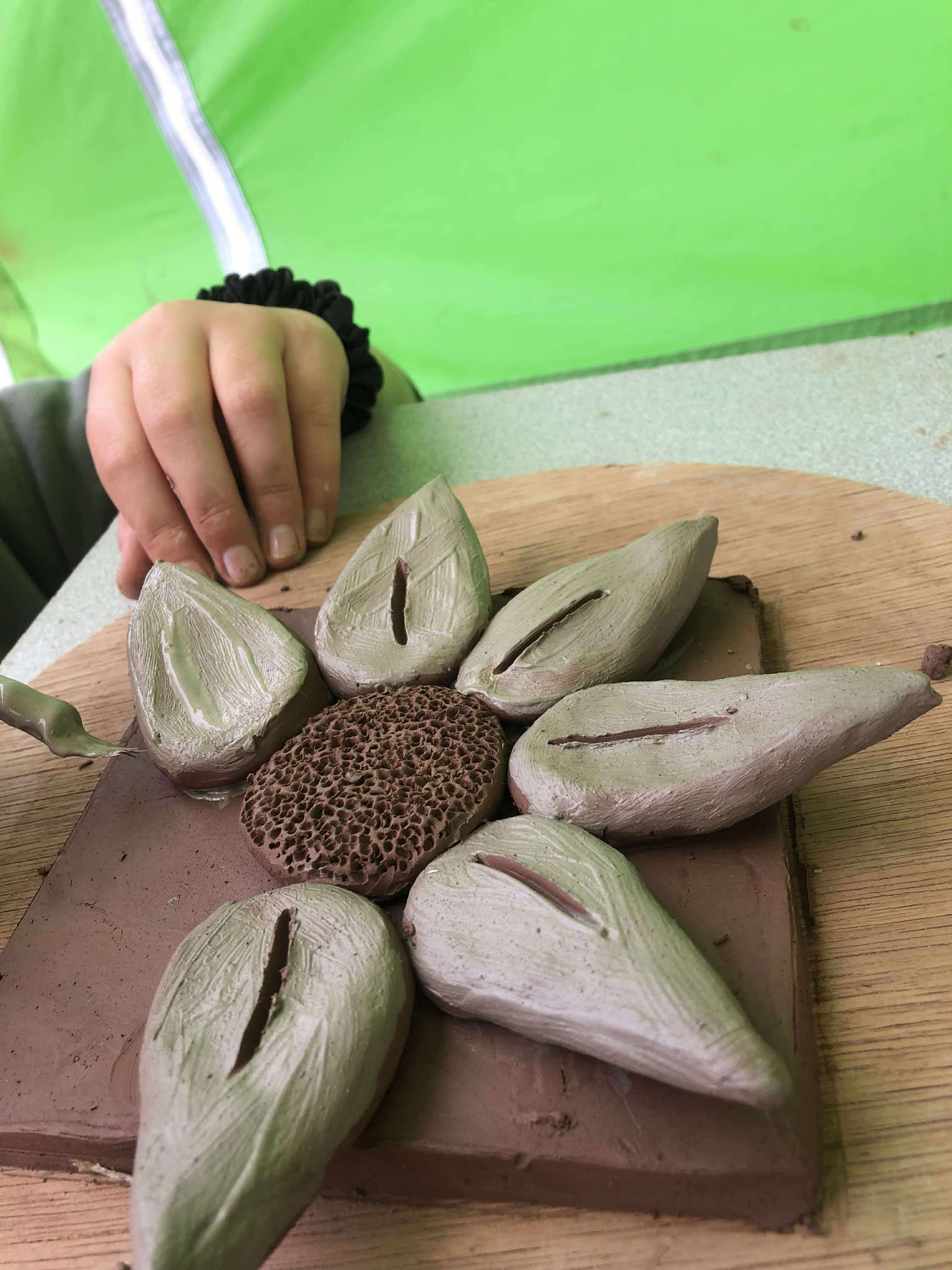 an unfinished modelled flower on a clay tile. flower has large x6 white petals. there is a gap where the final petal will be added. reminiscent of a sunflower
