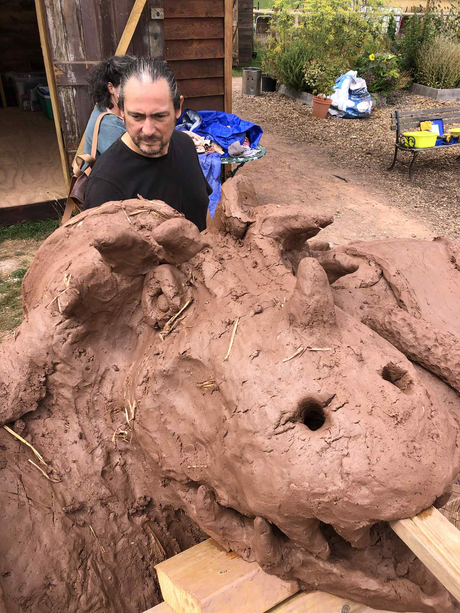 a male volunteer at growing local hereford helping model a giant dragon head made out of cob and sitting across a duel chambered cob pizza oven