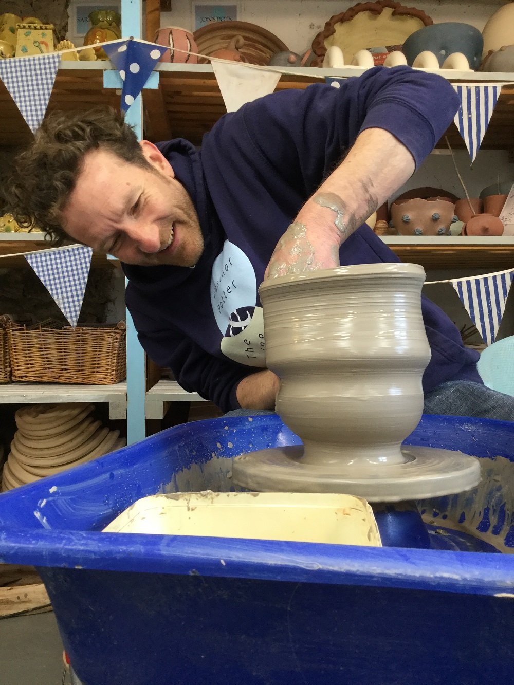 Jon Williams potter. Making a large grey pot on the potters wheel. He is leaning to the side and looking at the pot and he is smiling. He has one hand inside the pot and the other on the outside supporting. The shape is very fluid and undulating.