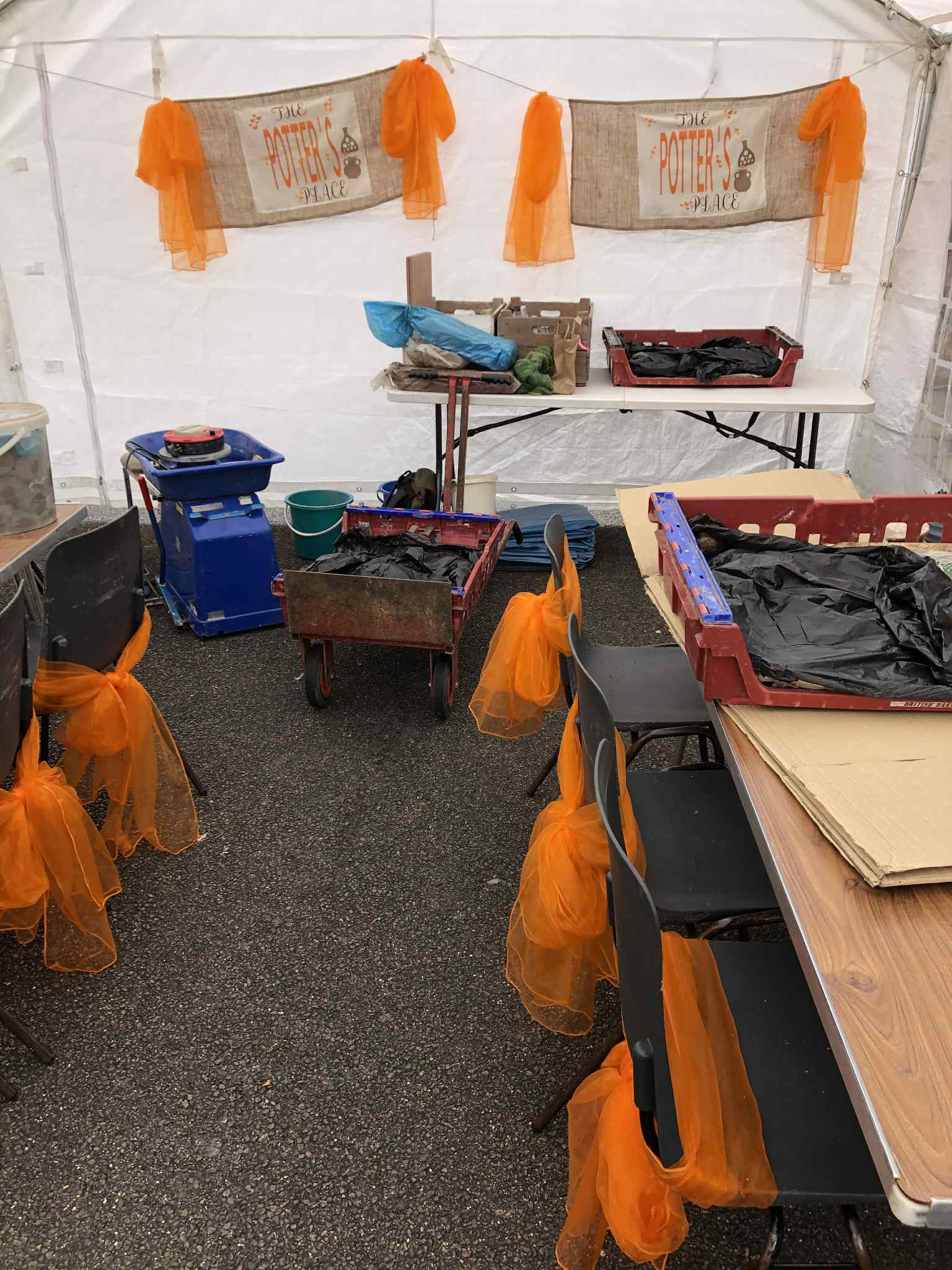 pop-up pottery studio in a white marquee containing tables chairs and a potter's wheel. There are two hand made banners hung from the end wall. Both display the text The Potter's Place