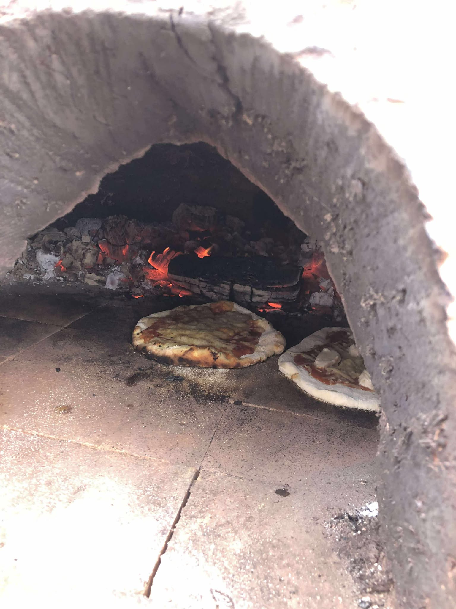 inside the pizza oven x2 freshly placed pizzas are cooking next to some glowing red embers