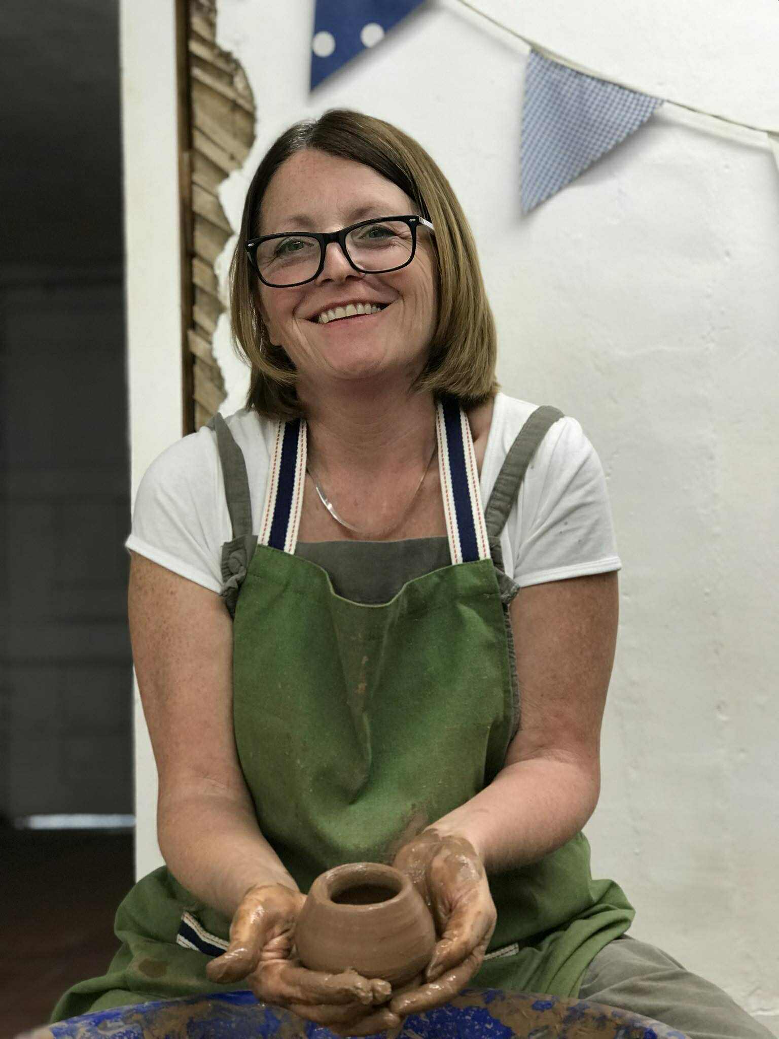 A lady wearing a green apron and wearing glasses. She is holding a freshly made pot which is nestled in both hands. She is smiling at the camera.