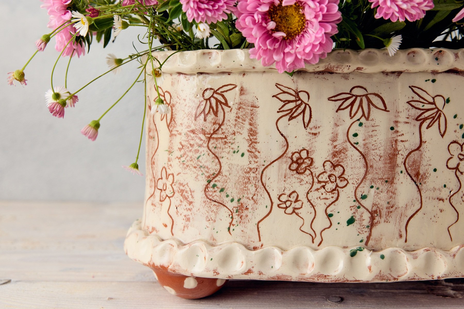 A detail of a front corner of a hand made, pottery flower brick made by Sarah Monk. It is white with etched flowers all over it. In the top there are a few bright pink flowers displayed in the top.