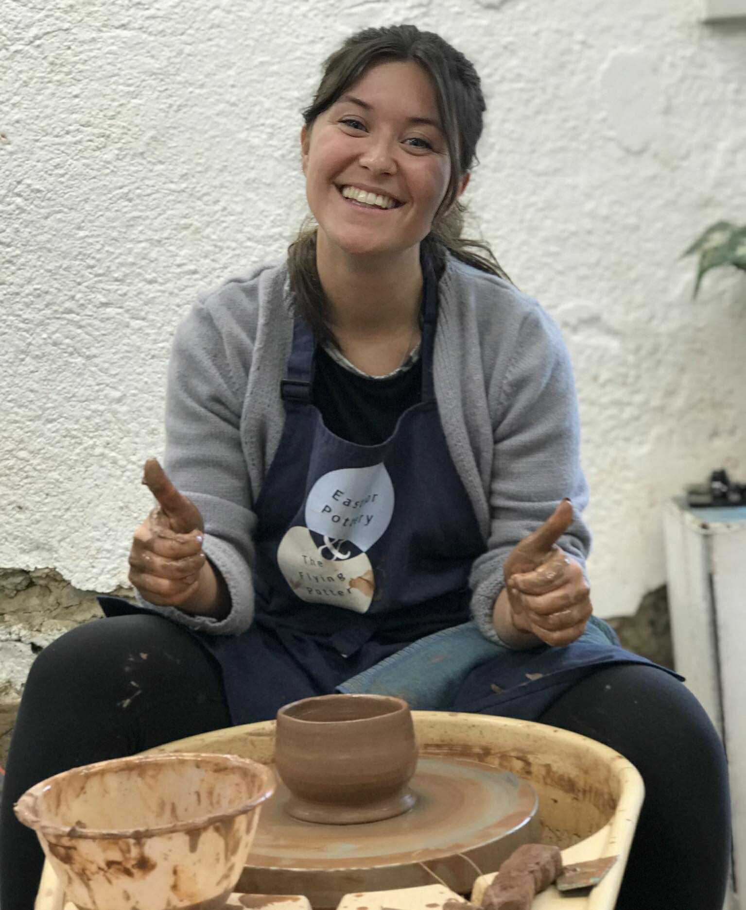 A young women sat at the potter's wheel wearing an Eastnor pottery apron. She has a freshly made bowl on her pottery wheel and is looking very happy. She is holding her thumbs up to suggest she is delighted with her pot.