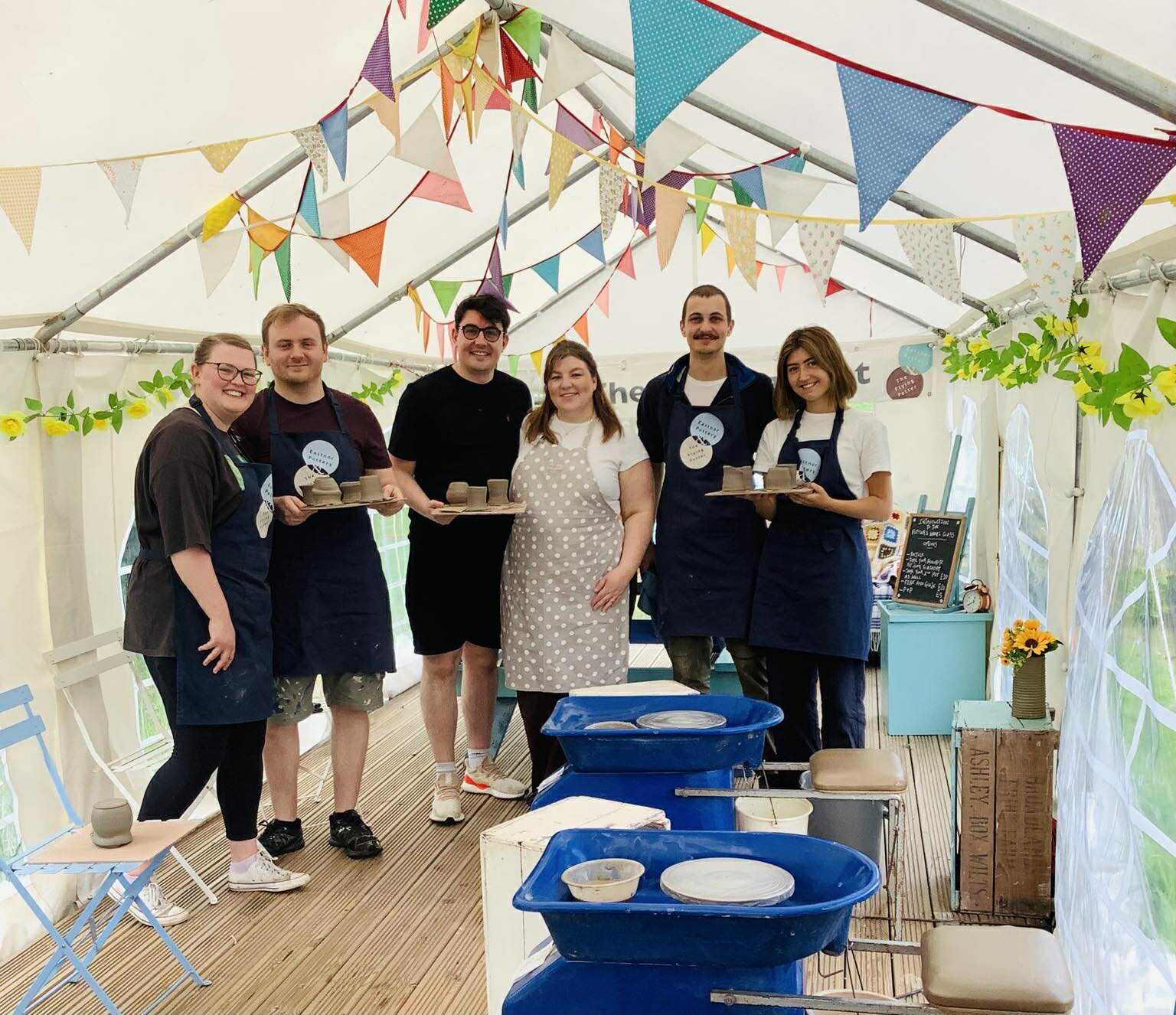 Four couple stood in a line in the potting tent marquee. The are holding up boards of freshly made pots, The tent is decorated with swaths of colourful bunting. The are stood in front of some pottery wheels.