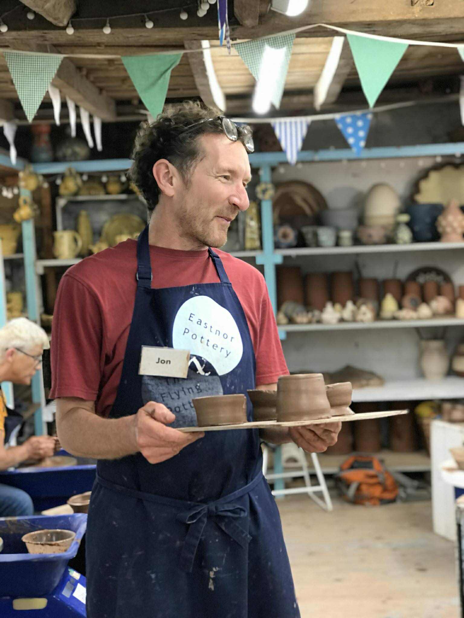 A man stood in the centre of the studio holding a board of freshly made pottery. He is wearing an Eastnor pottery apron and a name tag the say Jon.