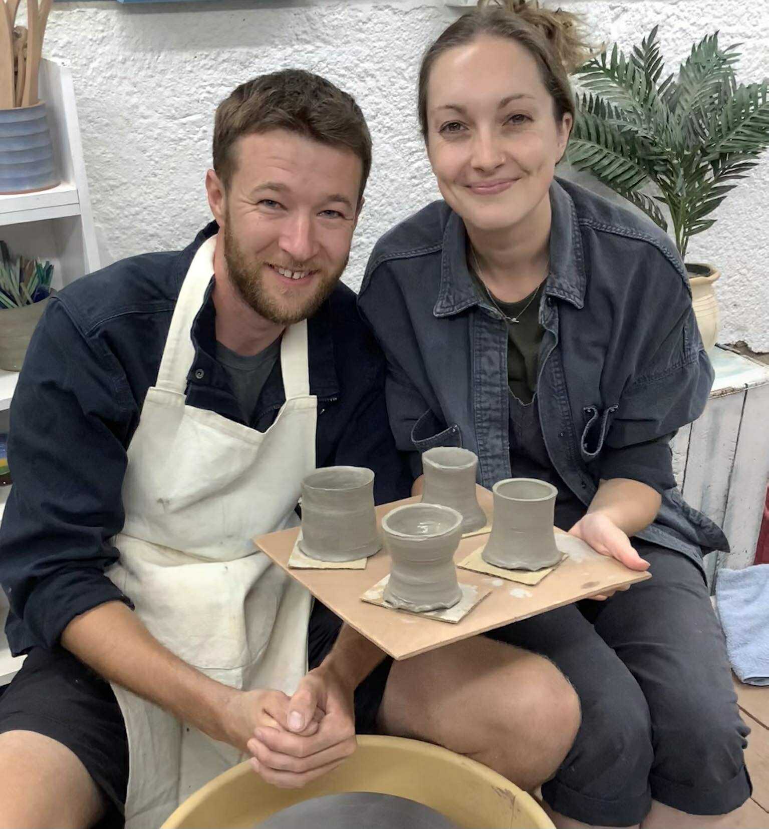 A young couple sat closely together at the potters wheel. They are holding up a board with 4 freshly made grey clay pots. They are smiling and looking at the camera
