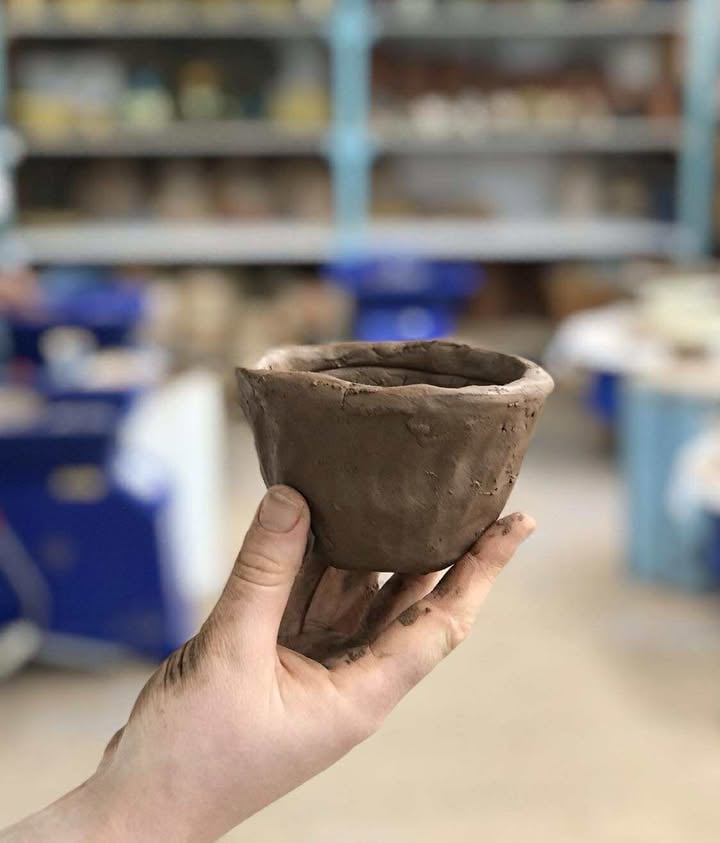 Hand shot holding a coil pot bowl up in the air, the background is of the studio shelves and pottery wheels but  the background is out of focus. 