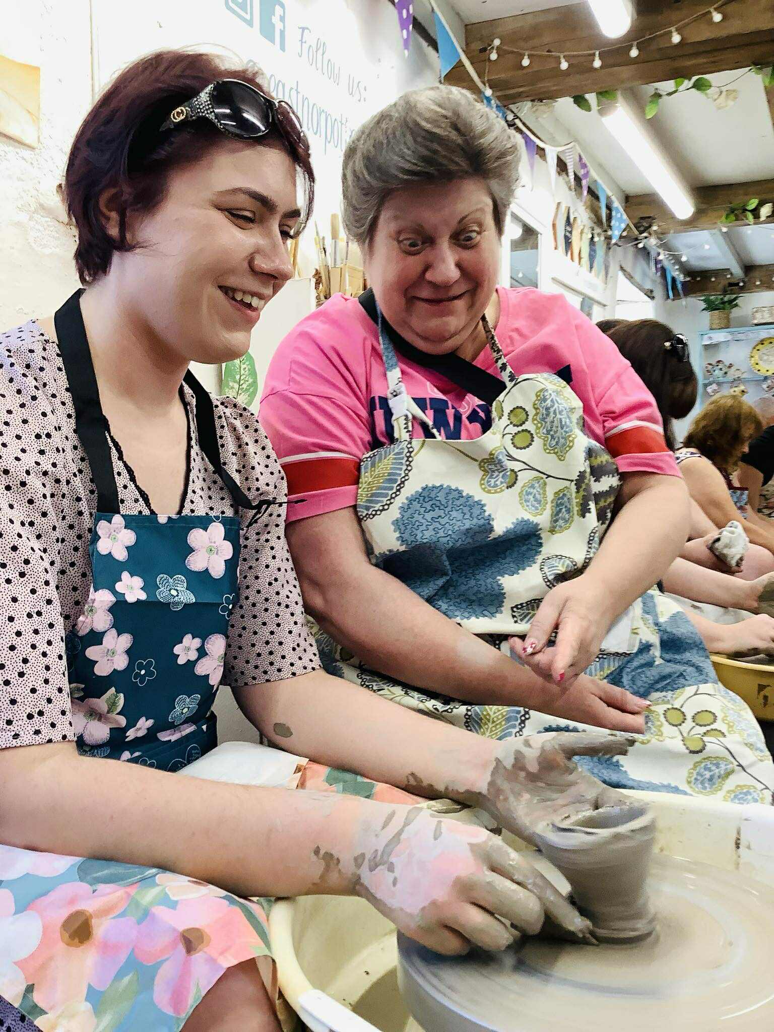 A mother and daughter are sat together at the wheel. The daughter has made a very woobly pot and she's attempting to remove it from the wheel. The mother is laughing at pointing at the pot.