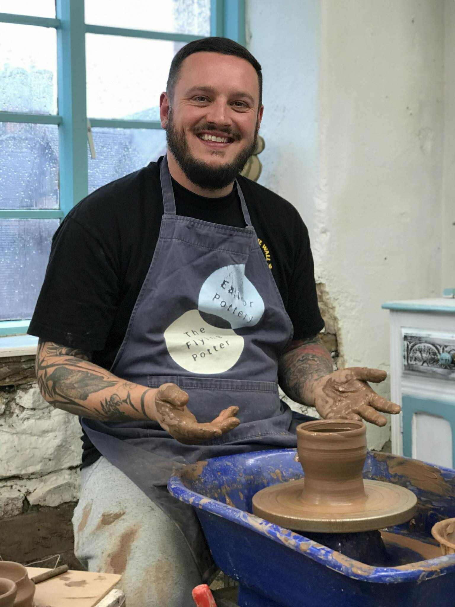 A man in his mid twenties, sat at the pottery wheel wearing an Eastnor pottery apron and smiling at the camera. He has a freshly made pot on the wheel and clay all over his hands
