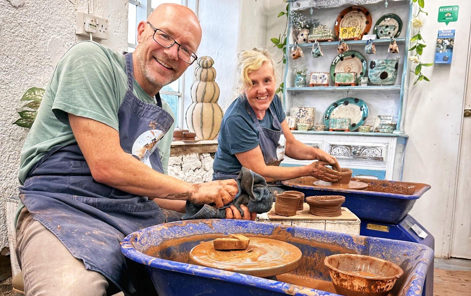 man and woman sat on their potter's wheel with brown clay in their hands and smiles on their faces looking directly into the camera. They are both wearing pottery aprons and surrounded by wet brown pots they have made during their pottery course at eastnor pottery herefordshire