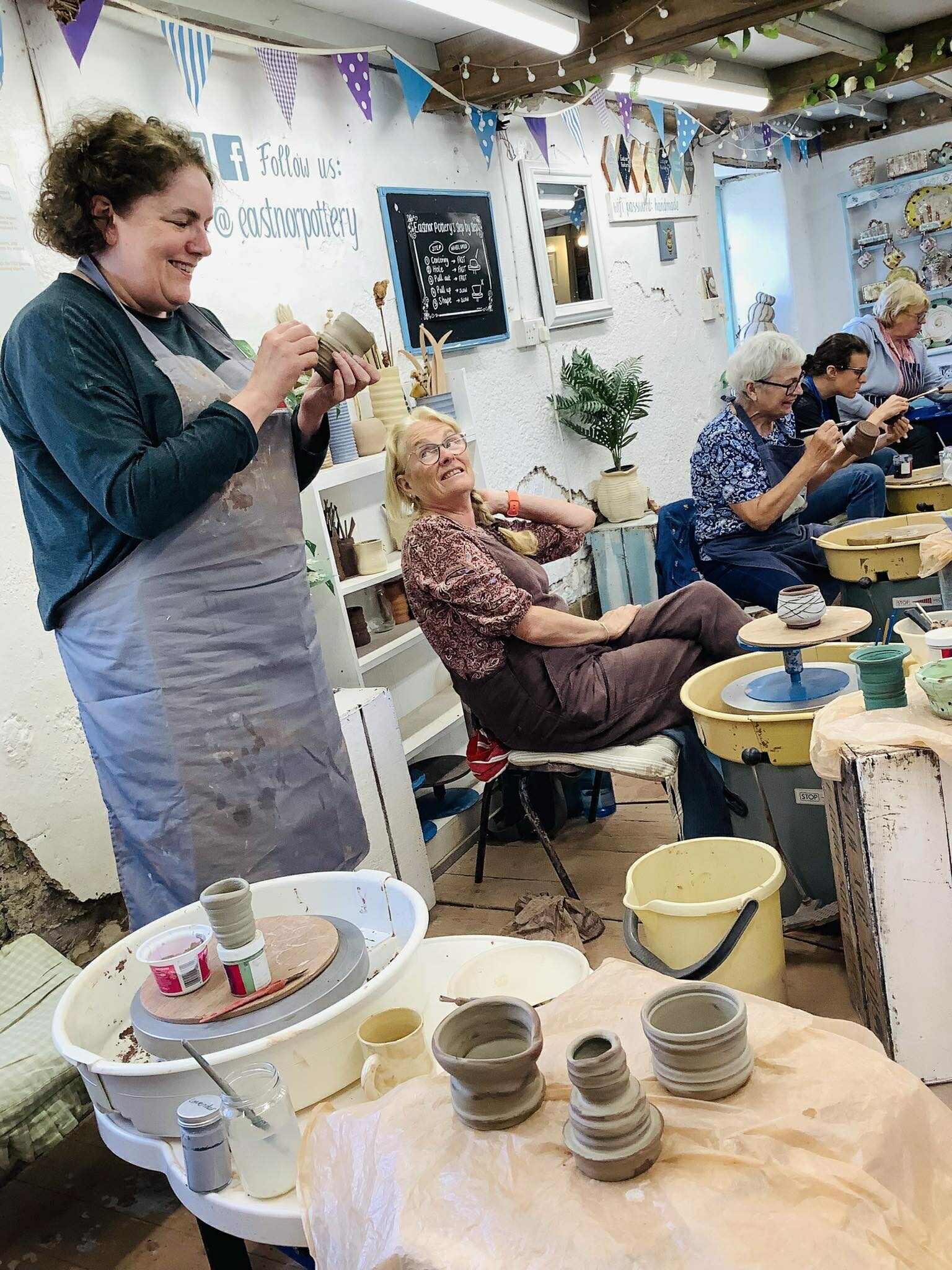 A lady in an apron is stood up holding a clay pot. She is decorating it whilst happily talking to another lady who is smiling and sat down on a chair at a potters wheel. there is a row of potters wheels and the people sat at them are busy decorating their pots with paints.
