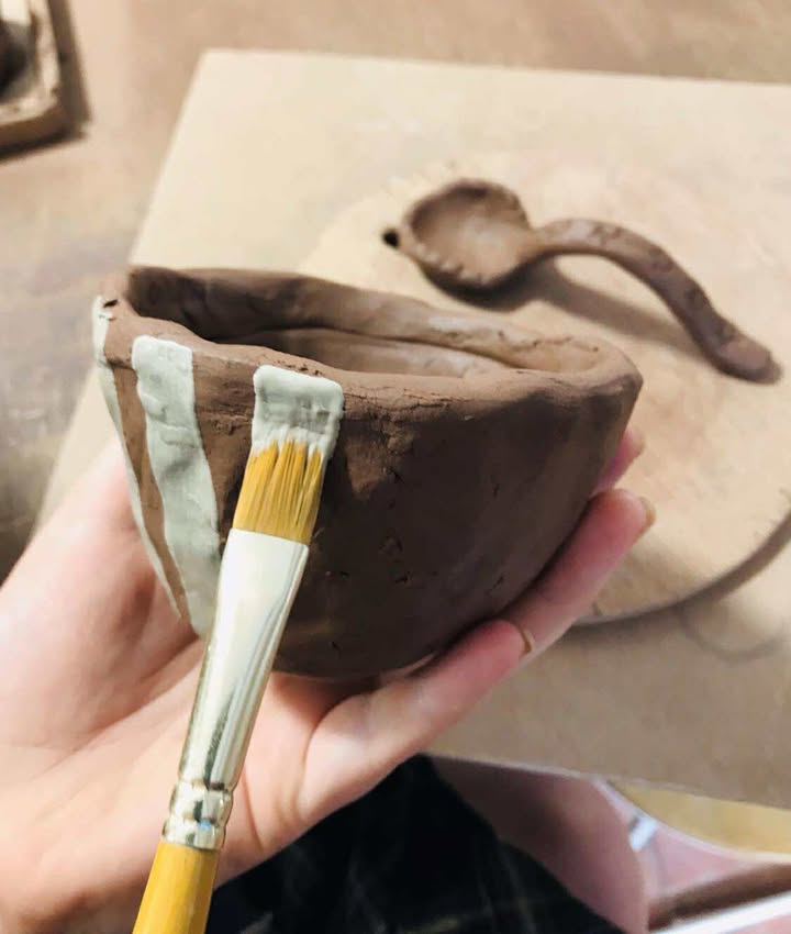 A close up of a hand holding a paint brush and painting on some white slip onto a terracotta bowl.