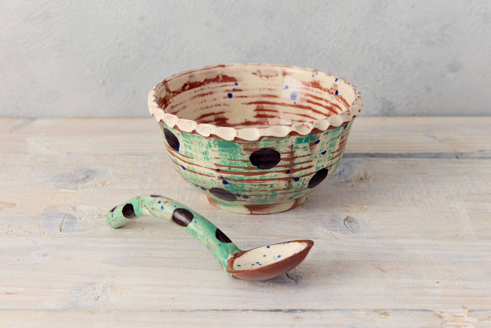 A pottery soup bowl with matching pottery spoon. The bowl is green with black polka dots, with a white pie crust rim. The spoon has a green handle with black dots. The spoon end is painted white. The bowl is made by Eastnor potter, Sarah Monk.