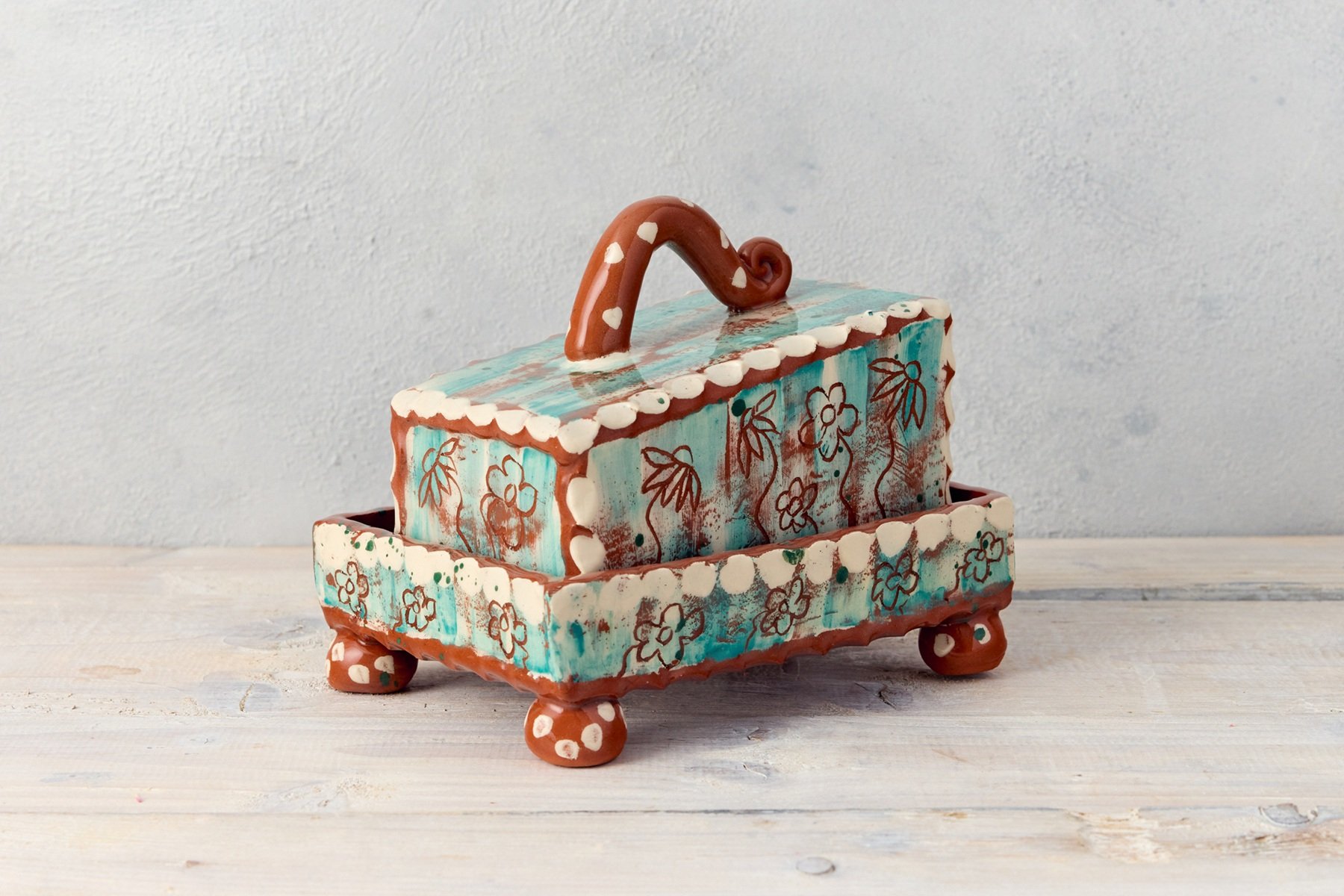 A teal slab built pottery butter dish with a slanted lid. The piece has etched flower motifs. It has a curly handle and the tray has little round feet with white dots.