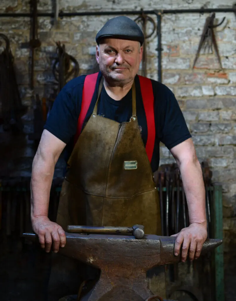 A portrait of artist blacksmith Andrew Findlay stood in his forge. He is wearing a back cap and leather apron. He is leaning on a large anvil with a hammer resting in between his hands. The backgound is an old stone wall with blacksmith tool hanging on it.