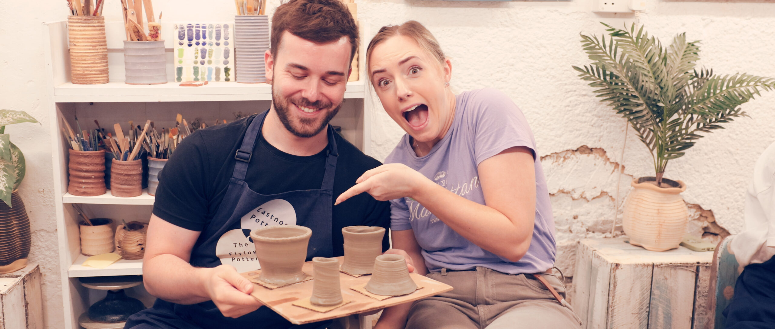 head and shoulders of young couple holding a board of four wet pots made on the potter's wheel at eastnor pottery. Both are smiling. the female is looking directly into the camera and pointing at her pot on the board. The male is looking down at his pot he made.