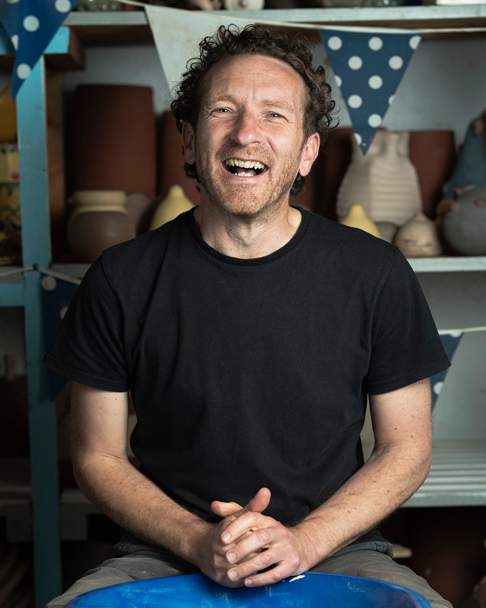 A portrait photo of Jon Williams. He is sat with his hands clasped together in front of him, leaning them on the edge of a blue pottery wheel. He is looking directly at the camera and laughing. He is wearing a black t-shirt and has curly brown hair. The are some shelves of ceramics behind with some hanging blue and white polka dot bunting