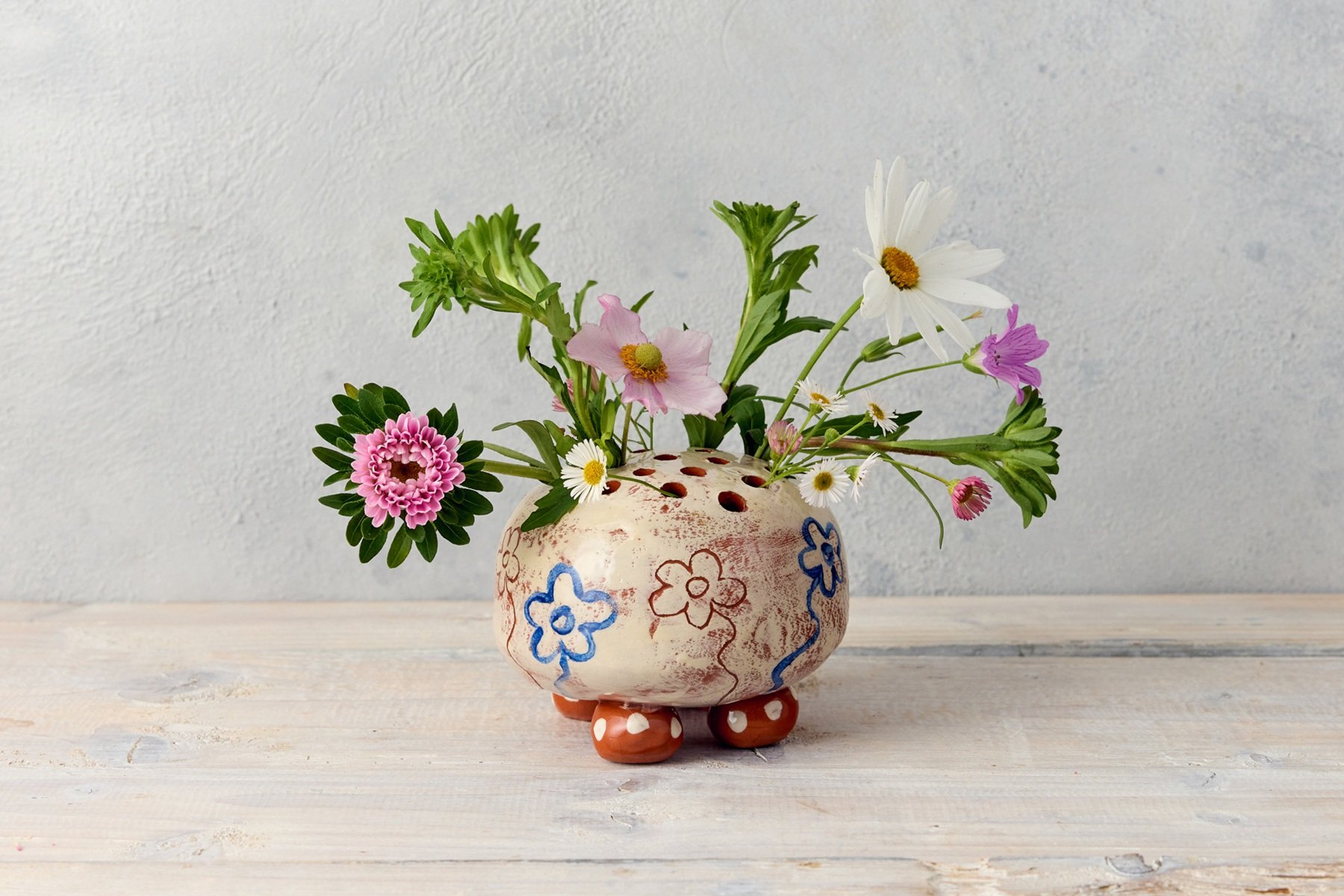 A round flower brick with holes in the top. It is white with simple painted flowers and sgraffito. The pot sits on 4 round dotty feet. The pot also contains an arrangement of fresh flowers, including daisies and cosmos.