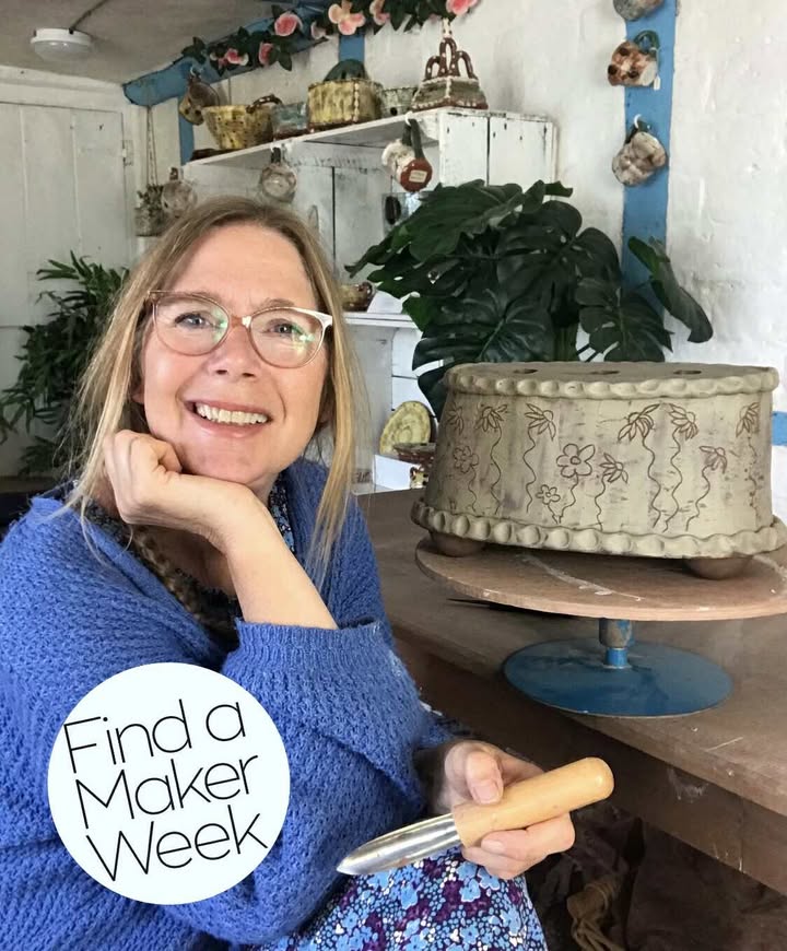 A portrait photo of Sarah Monk, smiling at the camera. She has blond hair and pink glasses and is wearing a blue cardigan. She is sat at her work table and is holding a metal pottery tool with a wooden handle. There is a large oval pot (approx. 30cms) on the table that she is currently working on.