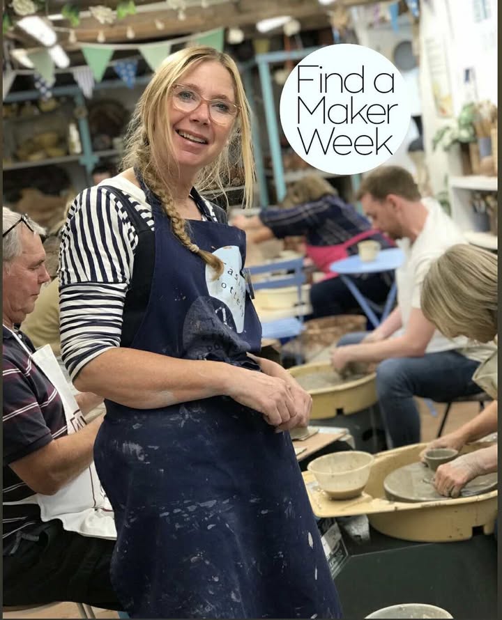 An action shot of Sarah Monk, wearing a stripey top and blue apron covered with clay. She is stood in the middle of the pottery studio smiling at the camera whilst teaching a potter's wheel class. The students are very engrossed in their work, taking pots off of the wheel.