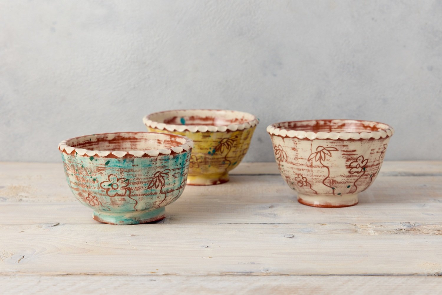 Three pottery bowls on a grey shelf, they are all different colours, from left to right: a teal one, a yellow one and a white one. They are decorated with brown etched flowers and the have white pie crust rims.