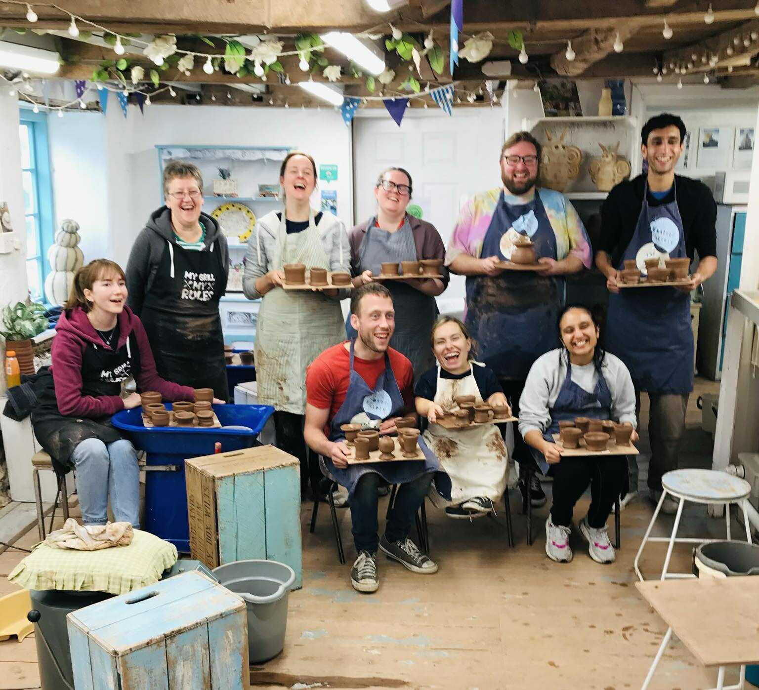 nine potter's wheel students attending a potter's wheel course at eastnor pottery herefordshire. There are five women and three men. Five are standing in a line at the back and four participants are sat on small chairs in the front. All are laughing and holding a board of brown freshly thrown terracotta pots they have made.