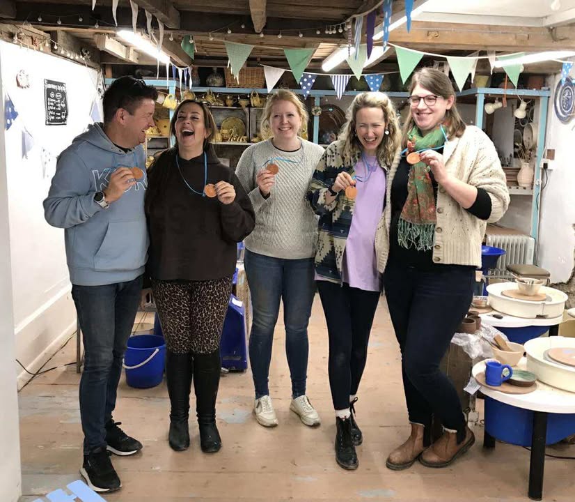 Five happy potters stood together in a line (1 man, 4 women) they are all laughing and they are proudly holding up their 'star potter' medals that are made in terracotta clay with a blue ribbon around each persons neck. They are holding the clay discs up to the camera.
